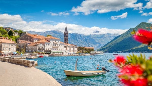 Boka Bay in Montenegro, a small and charming village on the shores of a fjord, with a stone church spire visible above the red roofs of the small buildings along the shore line. A small motor boat is in the foreground, with forested mountains in the background