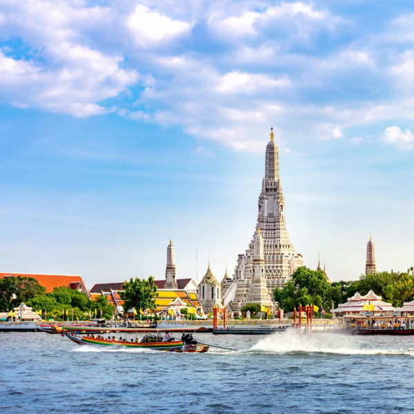 A traditional long tail boat filled with people in front of Wat Arun Temple, a dramatic spire-shaped white and gold temple