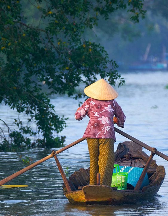 A person in traditional Vietnamese clothing standing on a shallow boat with two long oars, paddling through calm water