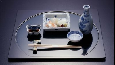 A rectangular plate laid out with Japanese food and drink items, including chopsticks on a chopstick resort, a blue and white pottery jug, a butterfly-shaped small pot filled with dark soy sauce, and a round bowl with delicate pieces of raw fish inside