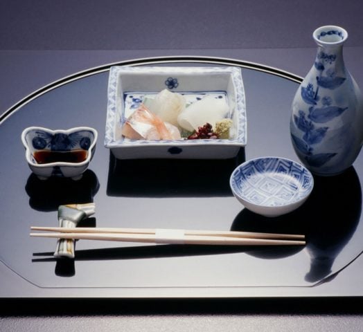 A rectangular plate laid out with Japanese food and drink items, including chopsticks on a chopstick resort, a blue and white pottery jug, a butterfly-shaped small pot filled with dark soy sauce, and a round bowl with delicate pieces of raw fish inside