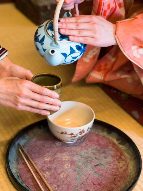 detail of two women in traditional kimono, kneeling on tatami preparing, pouring a cup of tea which is in the hands of one woman. They are in traditional Japanese old house on tatami.