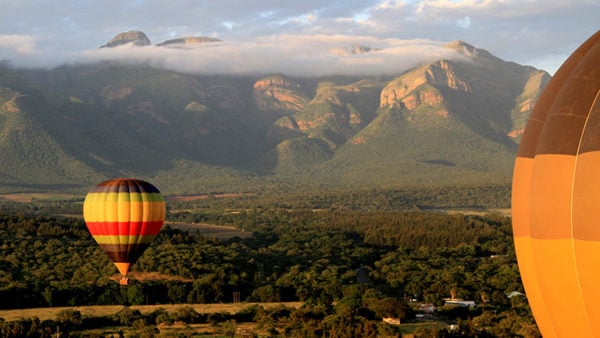 A rainbow striped hot air balloon soaring over fields and forests in South Africa with craggy mountains in the background