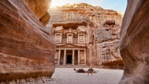 The temple-mausoleum of Al Khazneh in the ancient city of Petra in Jordan - a pinkish brown temple with columns craved out a tall craggy rock face. On the sand in front of the temple lies two camels with blankets and saddles, alongside a standing dog