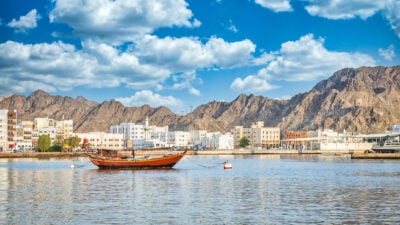 Old traditional sailboat anchored at Muttrah Corniche. The old city with white and sand coloured cube-shaped buildings and mountains are in the background. From Muscat, Oman.