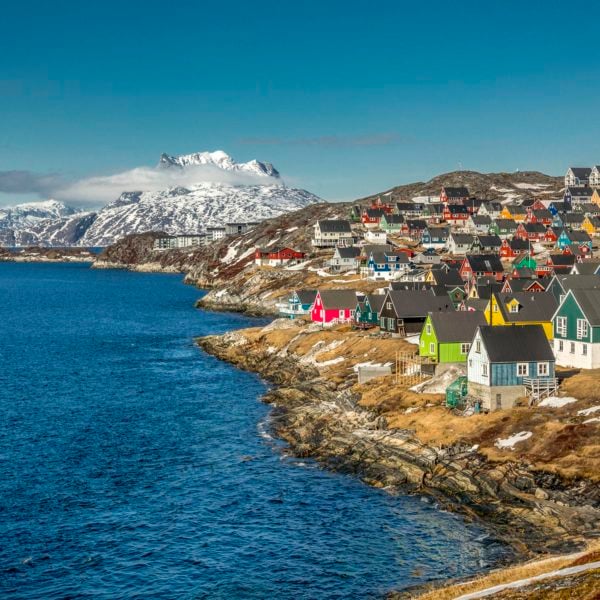 The town of Nuuk, Greenland, with rainbow painted traditional hut-style houses on a rocky shoreline, with snowy mountains in the background