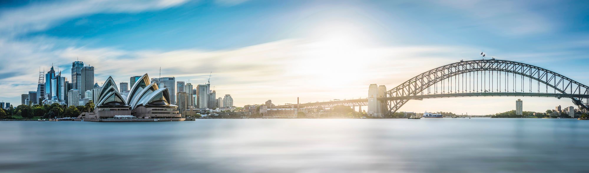 Panorama of Sydney skyline on a sunny autumn afternoon, showing the large curved iron shape of the harbour bridge, the sails of the opera house, and the skyscrapers of the city.