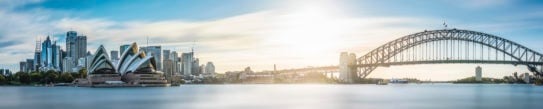 Panorama of Sydney skyline on a sunny autumn afternoon, showing the large curved iron shape of the harbour bridge, the sails of the opera house, and the skyscrapers of the city.