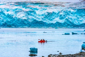 Two people wearing bright red coats and warm hats in a red kayak paddling past a large glacier and amongst icebergs